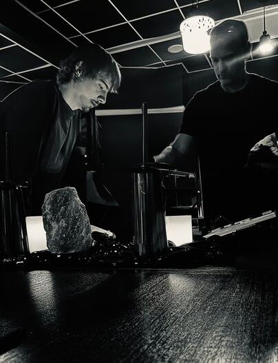escape room image. Two men in a dark studio inspect equipment on a wooden counter, with a rough rock centerpiece under overhead lights.